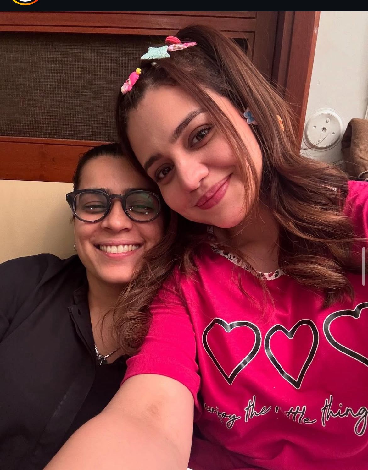 Two women smiling for a selfie, one wearing glasses and a black shirt, the other in a red T-shirt with three heart outlines and the text 'Enjoy the little things,' with colorful hair clips in her hair."